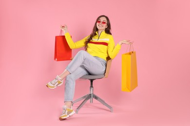 Happy woman in stylish sunglasses holding colorful shopping bags on armchair against pink background