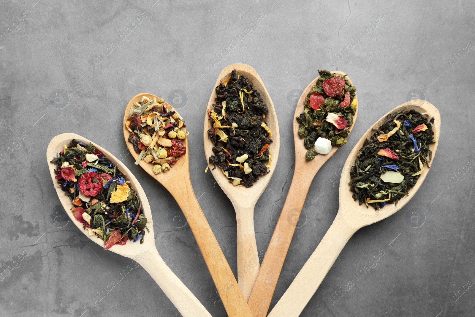 Photo of Different kinds of dry herbal tea in wooden spoons on light grey table, flat lay