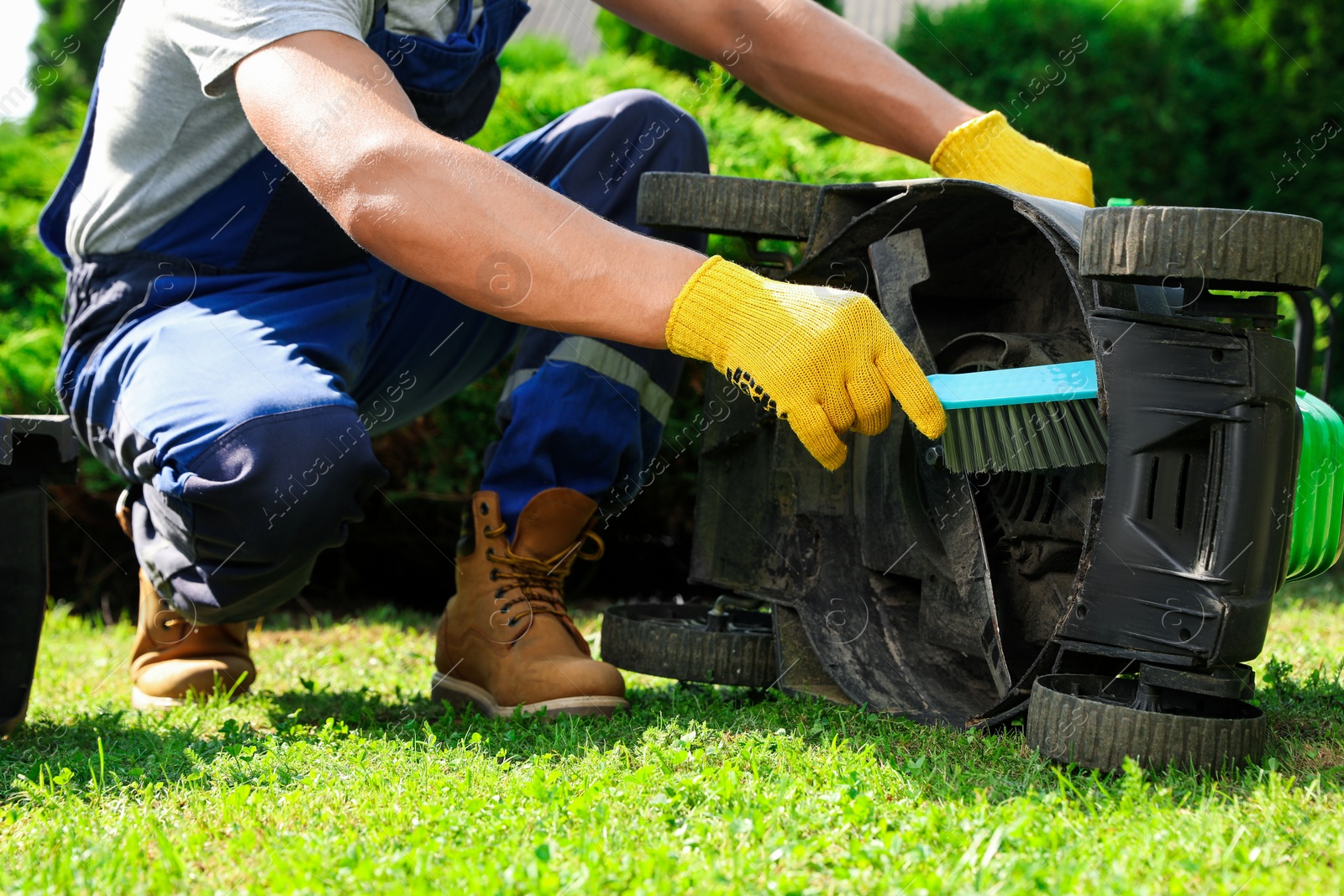 Photo of Young man cleaning lawn mower with brush in garden, closeup