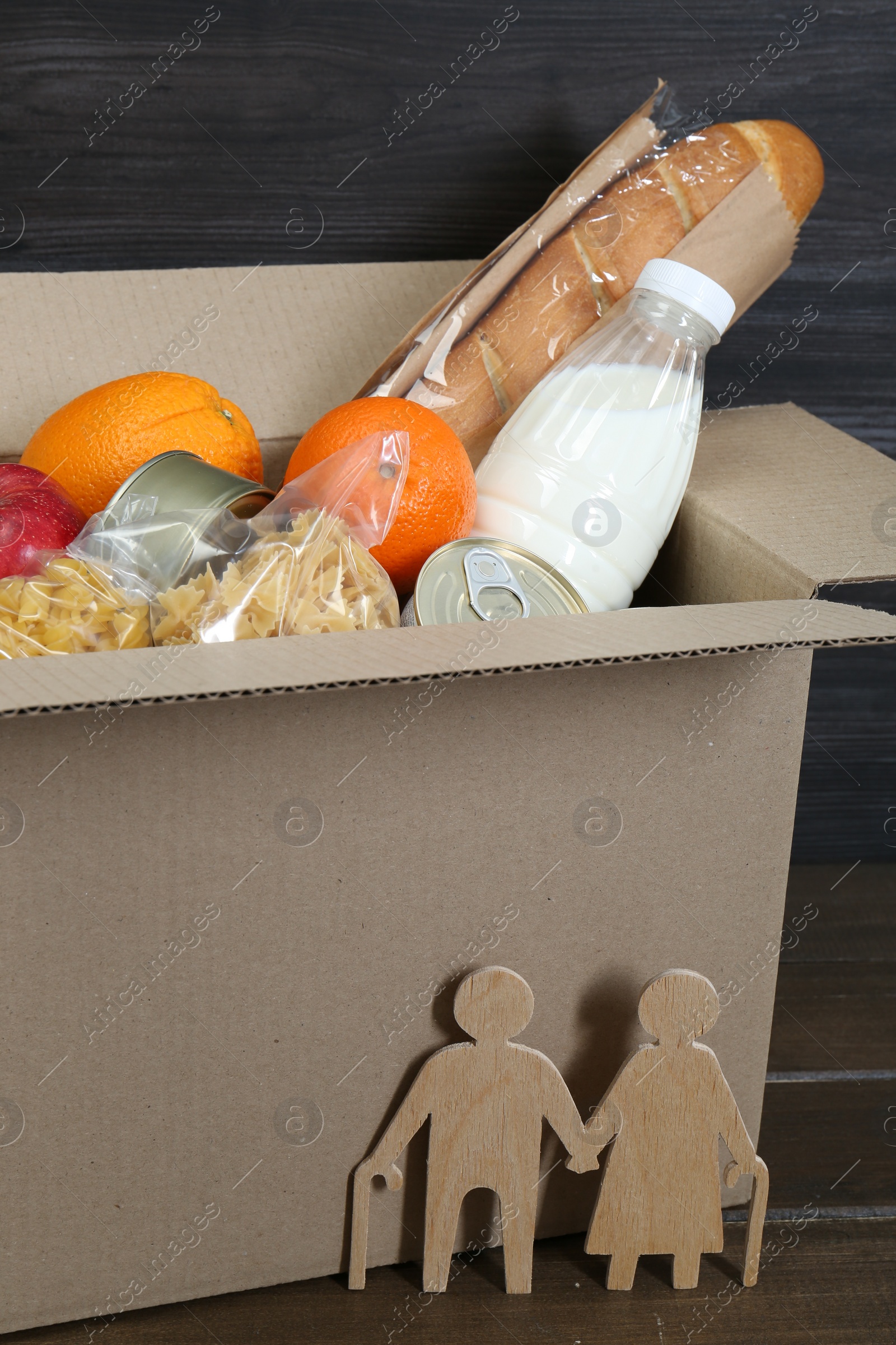 Photo of Humanitarian aid for elderly people. Cardboard box with donation food and figures of couple on wooden table, closeup