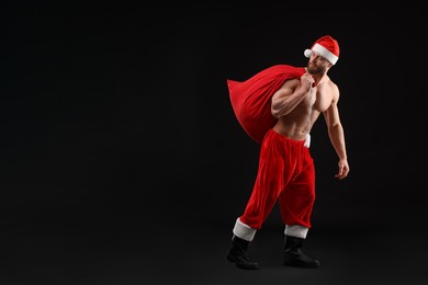 Muscular young man in Santa hat holding bag with presents on black background, space for text