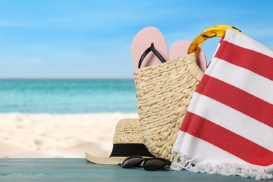 Beach bag with towel, flip flops, hat and sunglasses on light blue wooden surface near seashore. Space for text