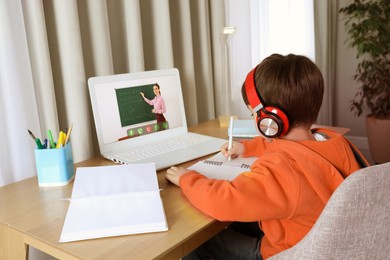 Image of E-learning. Little boy taking notes during online lesson at wooden table indoors