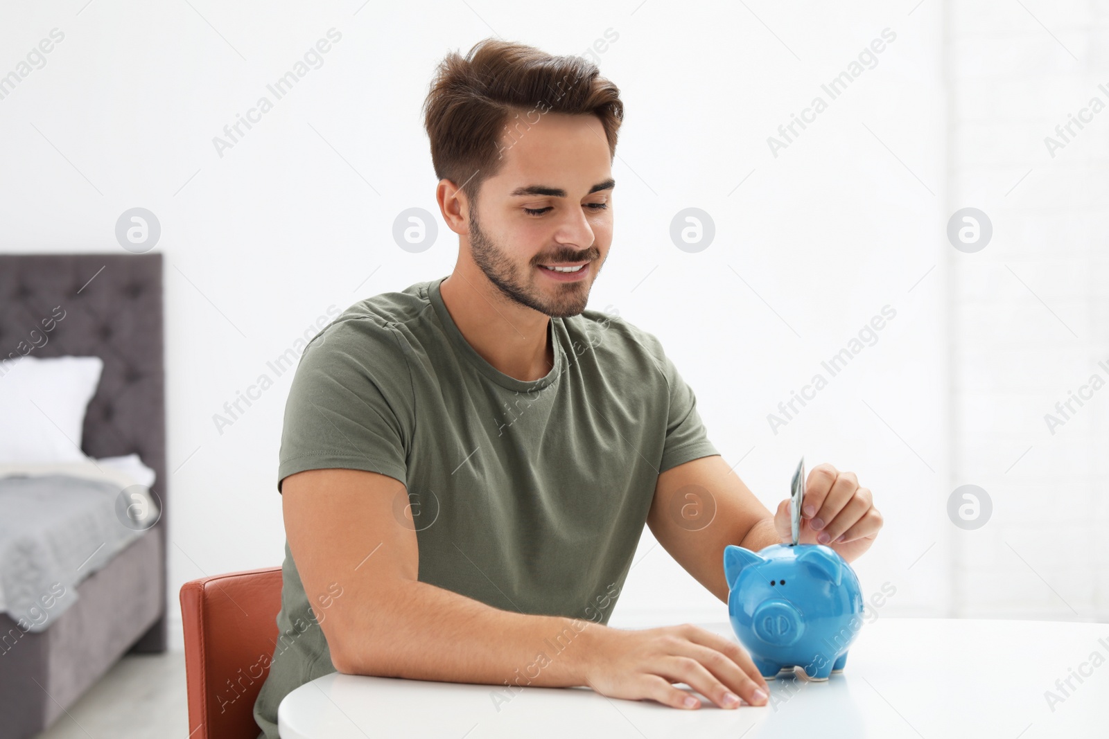 Photo of Young man putting money into piggy bank at table indoors