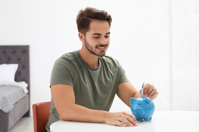 Young man putting money into piggy bank at table indoors