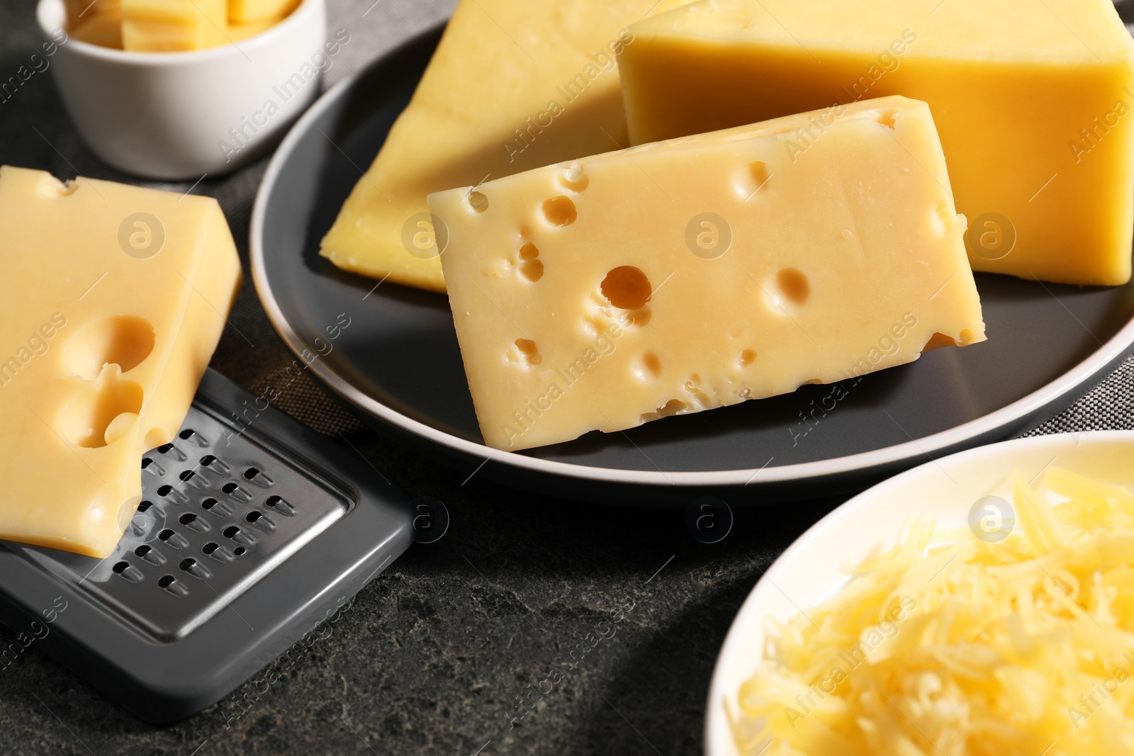 Photo of Grated, whole pieces of cheese and grater on dark textured table, closeup