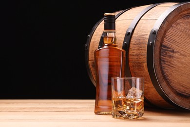 Photo of Whiskey with ice cubes in glass, bottle and barrel on wooden table against black background, space for text