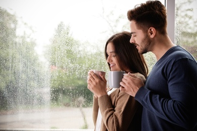 Happy young couple with cups near window indoors on rainy day