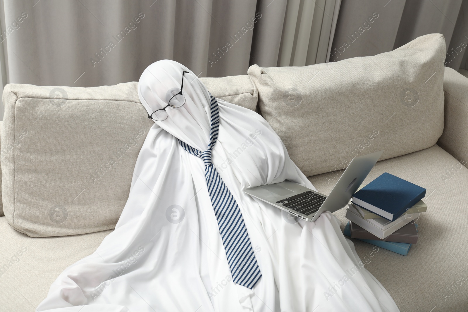Photo of Overworked ghost. Man in white sheet with laptop and books on sofa at home