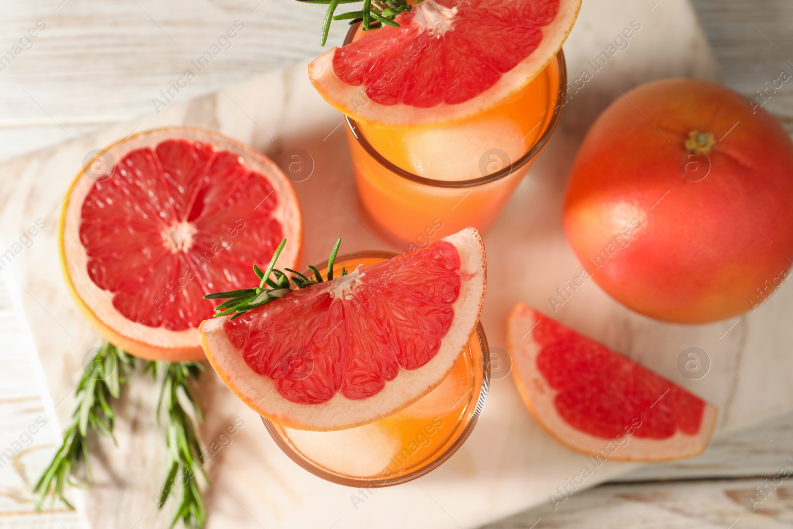 Photo of Tasty grapefruit drink with ice in glasses, rosemary and fresh fruits on light wooden table, flat lay