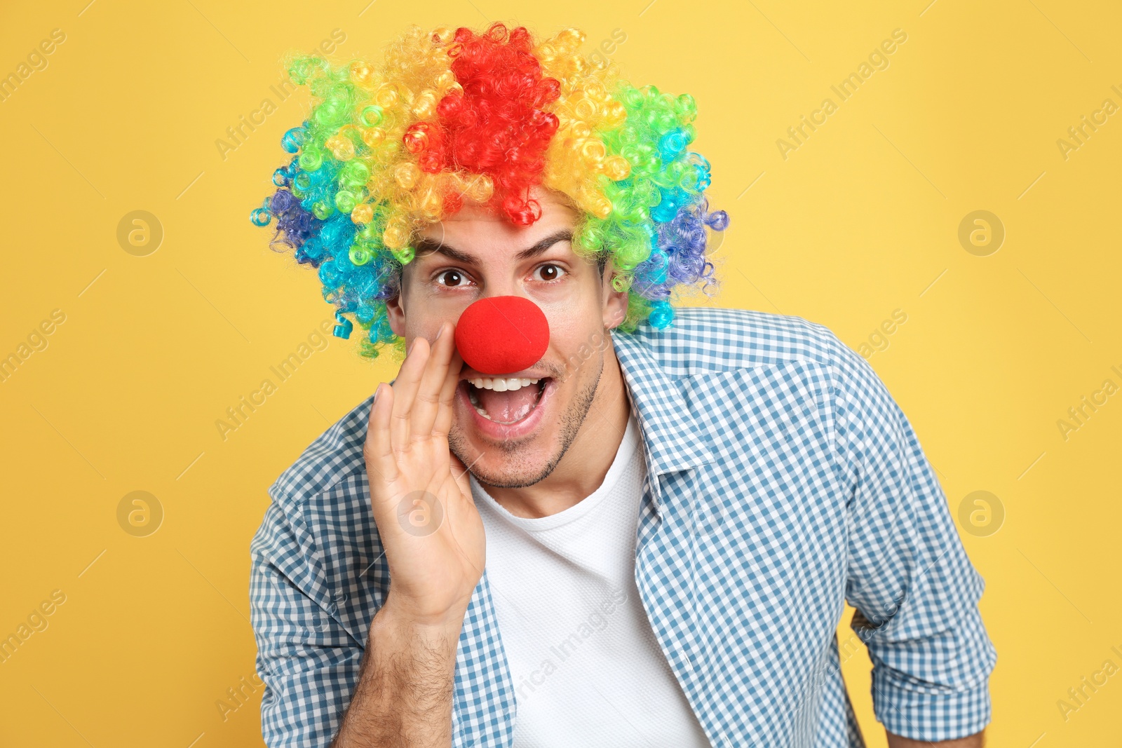 Photo of Funny man with clown nose and rainbow wig on yellow background. April fool's day