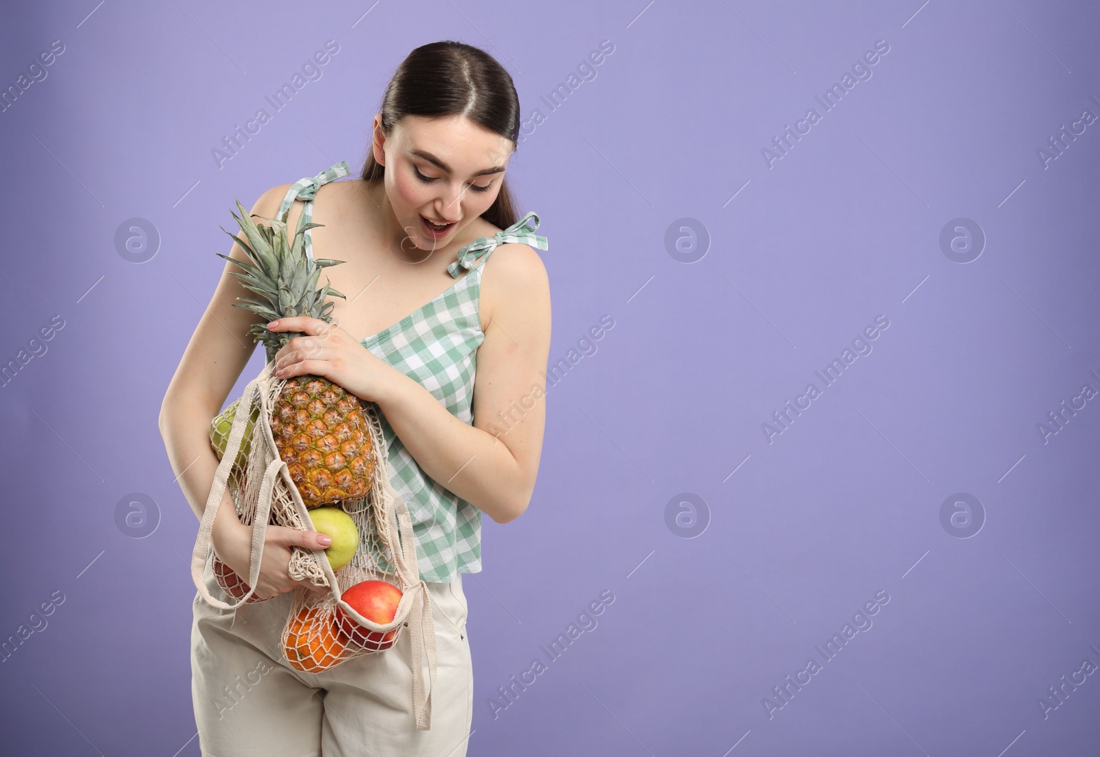 Photo of Surprised woman with string bag of fresh fruits on violet background, space for text