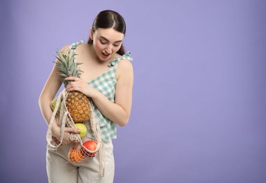 Surprised woman with string bag of fresh fruits on violet background, space for text