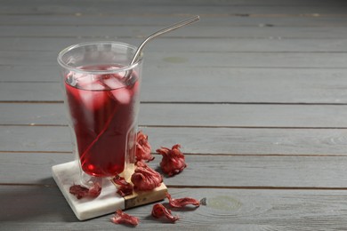 Refreshing hibiscus tea with ice cubes in glass and roselle petals on grey wooden table. Space for text