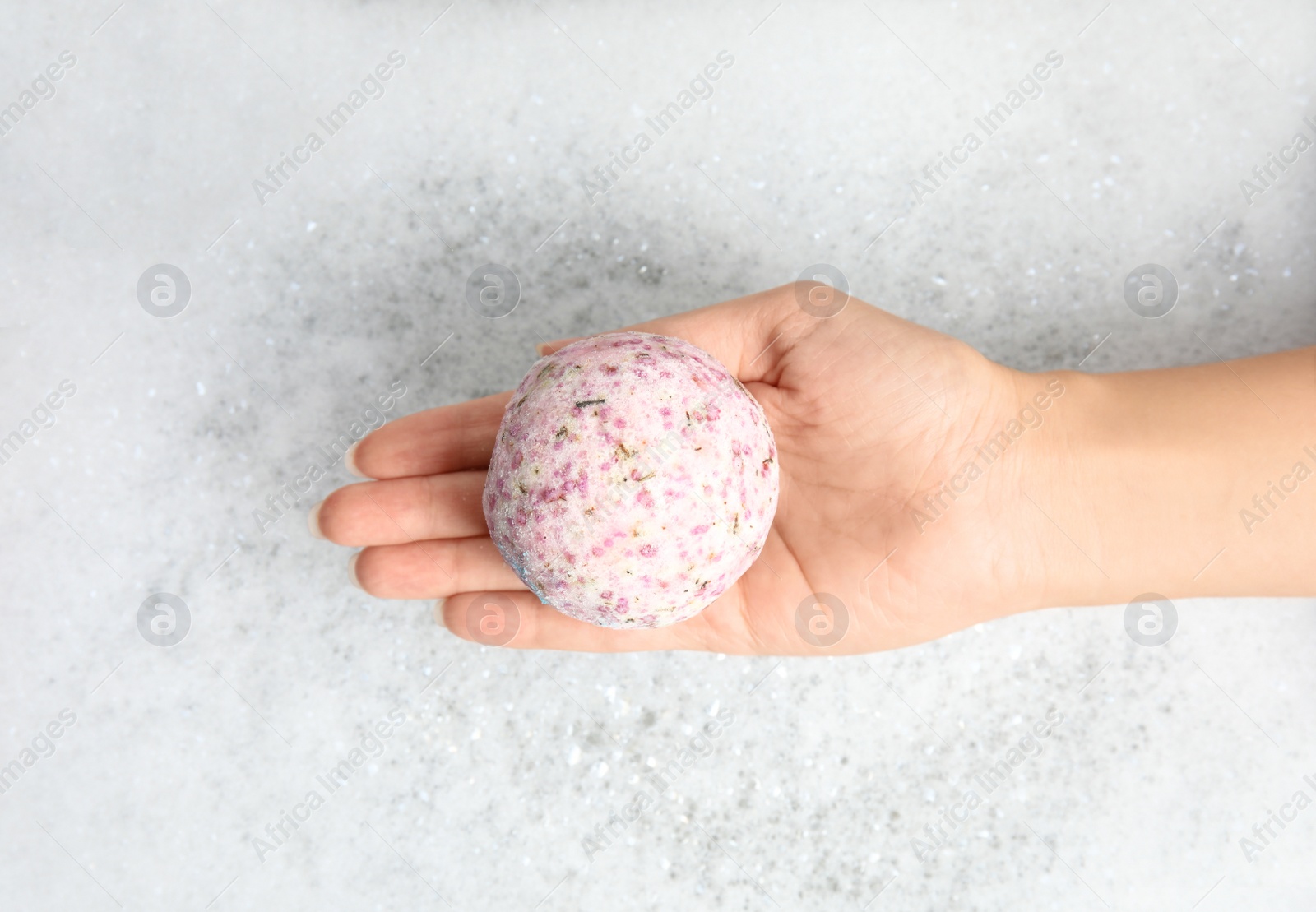 Photo of Woman holding color bath bomb over foam, top view