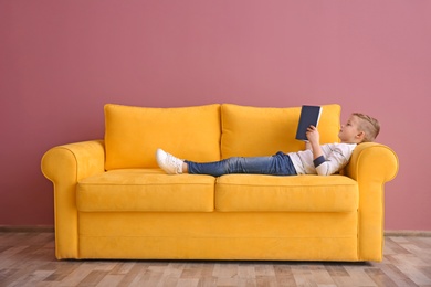 Cute little boy lying on sofa and reading book, indoors