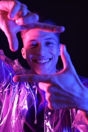 Young man making camera with hands on dark background in neon lights