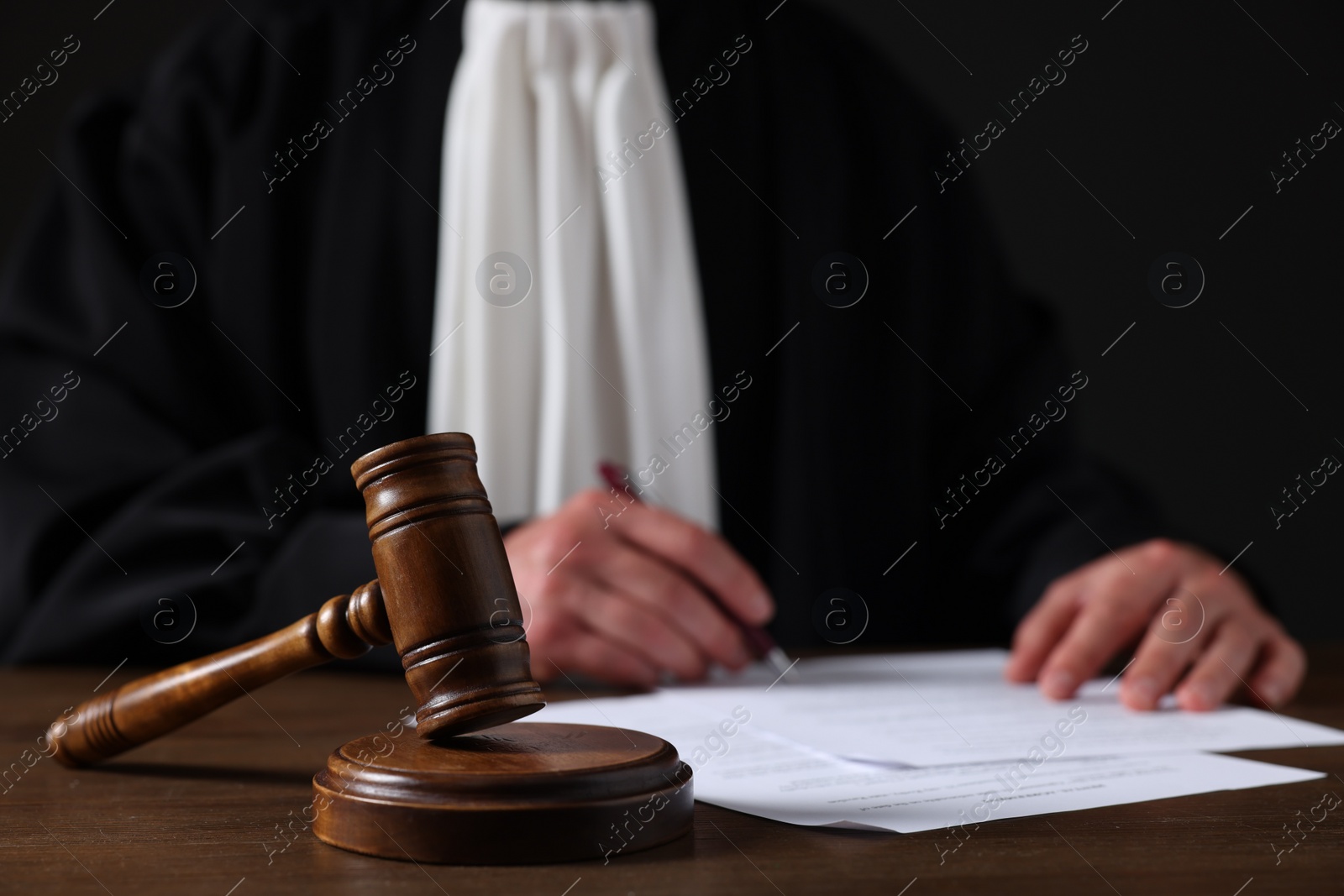 Photo of Judge with gavel writing in papers at wooden table against black background, closeup