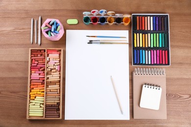 Photo of Blank sheet of paper, colorful chalk pastels and other drawing tools on wooden table, flat lay. Modern artist's workplace