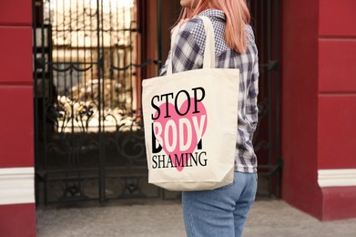 Image of Woman holding bag with words Stop Body Shaming near building outdoors, closeup