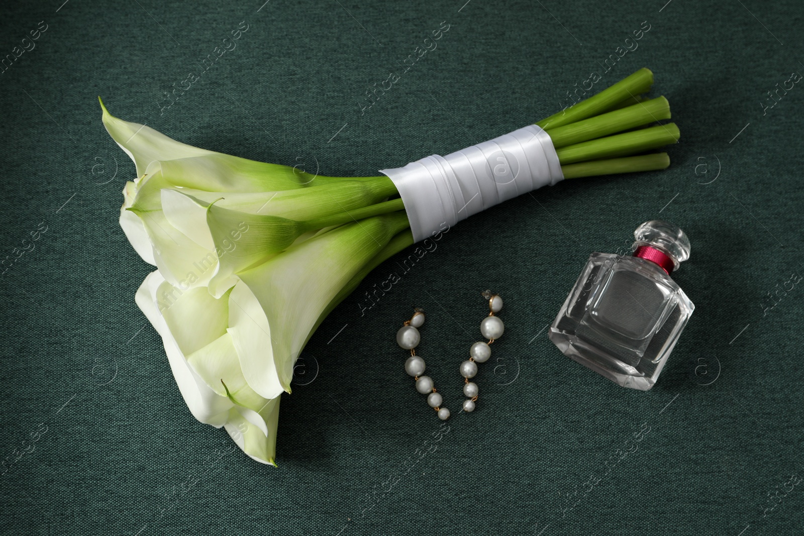 Photo of Beautiful calla lily flowers tied with ribbon, bottle of perfume and earrings on green fabric, above view