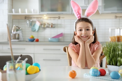 Photo of Cute little girl with bunny ears headband and painted Easter eggs sitting at table in kitchen, space for text