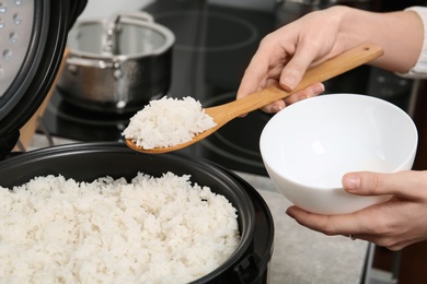 Photo of Woman putting boiled rice into bowl from cooker in kitchen, closeup