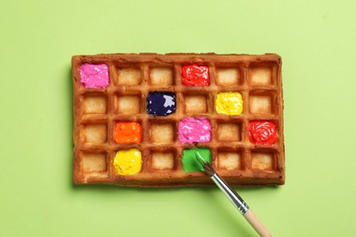 Photo of Waffle with different paints and brush on light green background, top view