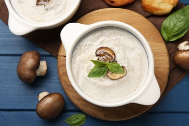 Photo of Delicious mushroom soup in ceramic pots and fresh ingredients on blue wooden table, flat lay