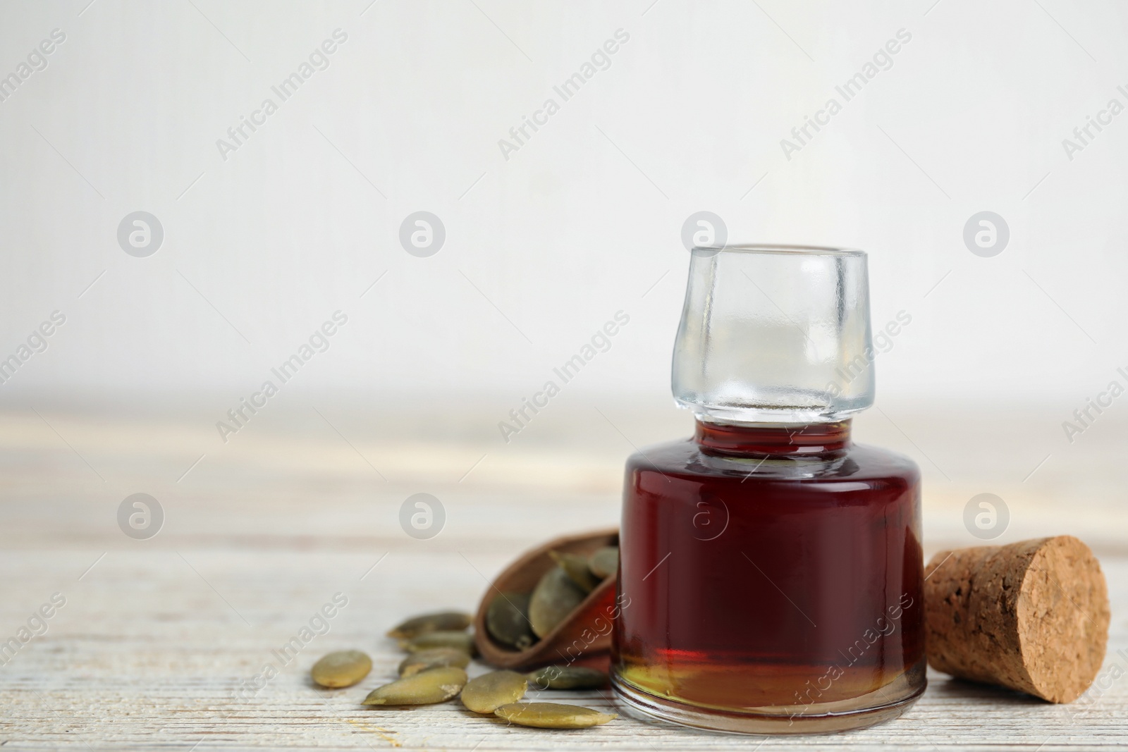 Photo of Glass bottle of oil and scoop with pumpkin seeds on white wooden table, closeup. Space for text