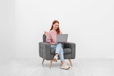 Happy young woman having video chat via laptop while sitting in armchair near white wall indoors