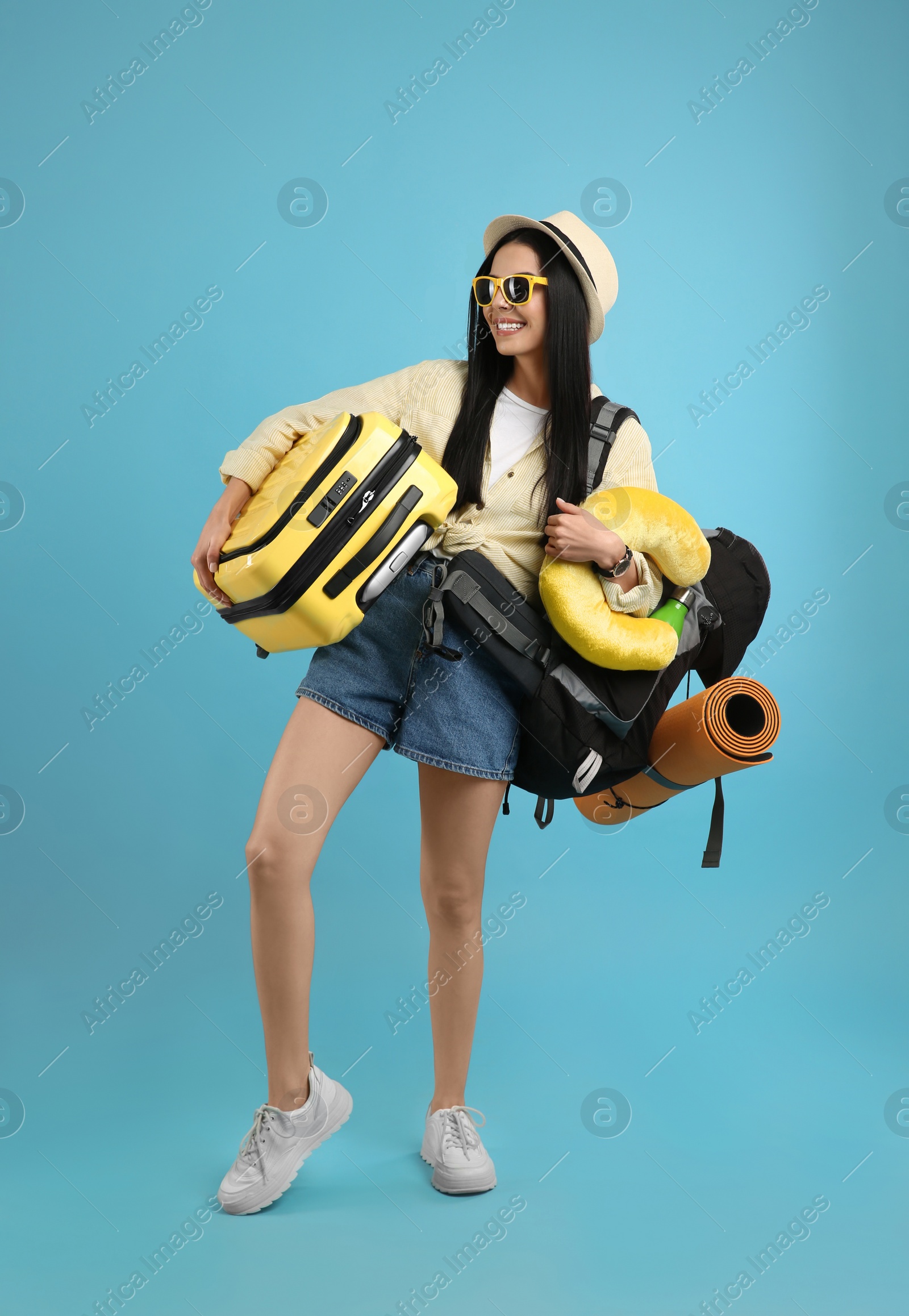 Photo of Happy female tourist with backpack, suitcase and travel pillow on light blue background