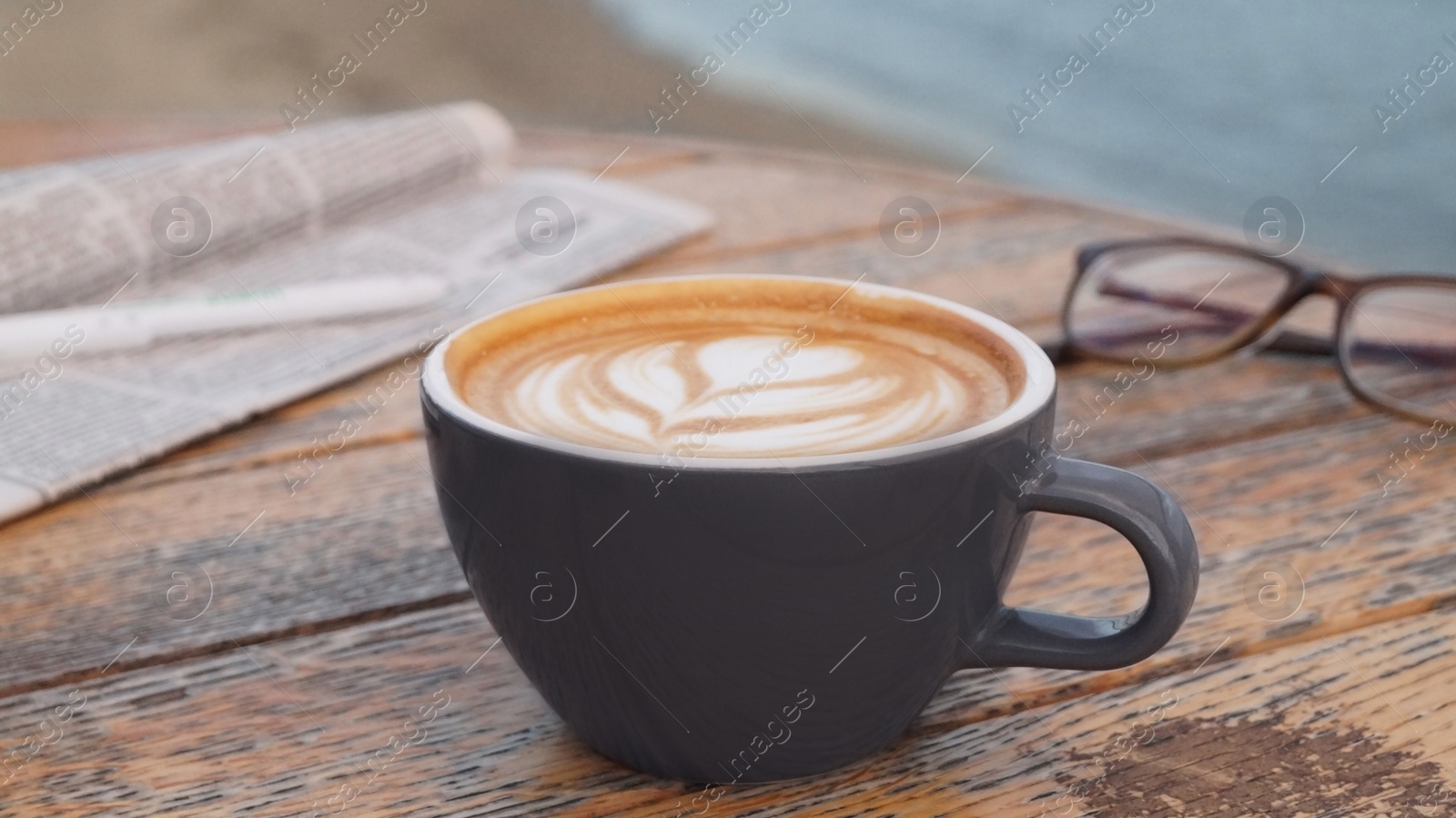 Photo of Cup of delicious coffee, eyeglasses and newspaper on wooden table, closeup
