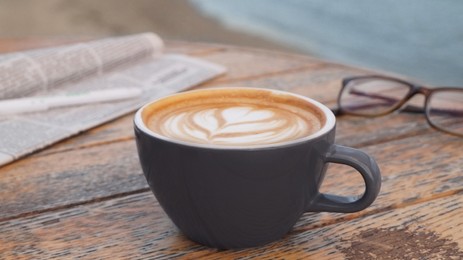 Photo of Cup of delicious coffee, eyeglasses and newspaper on wooden table, closeup