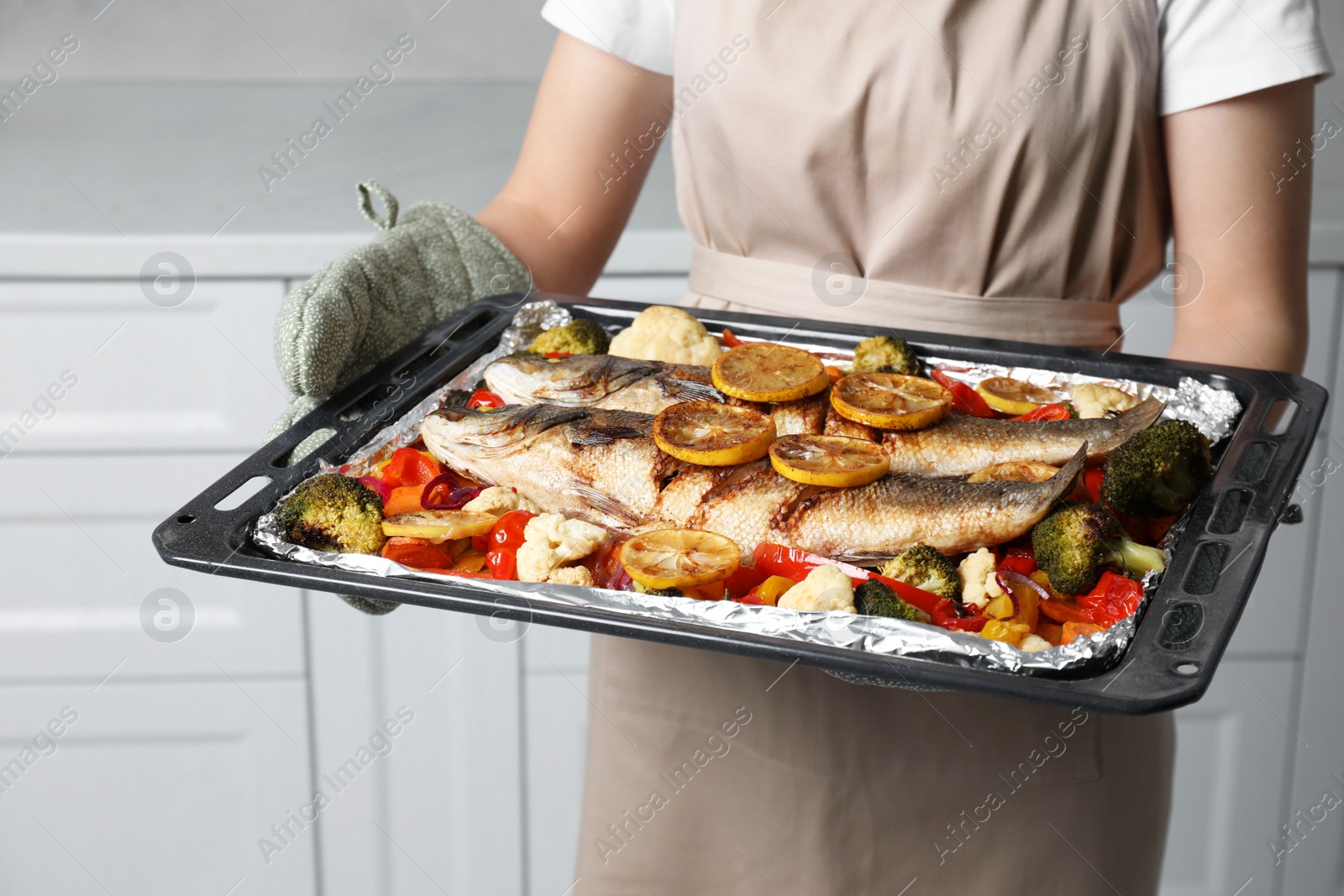 Photo of Woman holding baking tray with sea bass fish and vegetables in kitchen, closeup