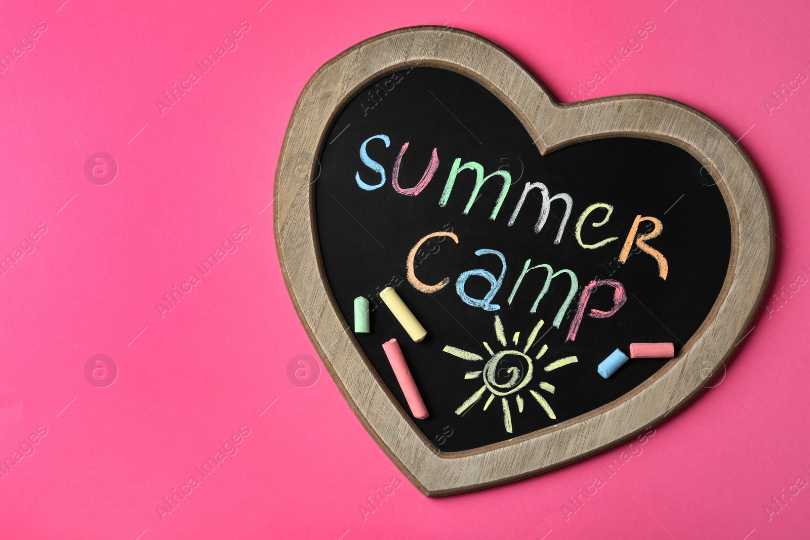 Photo of Heart shaped blackboard with text SUMMER CAMP, drawing and chalk sticks on color background, top view. Space for design