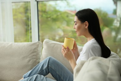 Young woman with cup of coffee relaxing on sofa at home, space for text