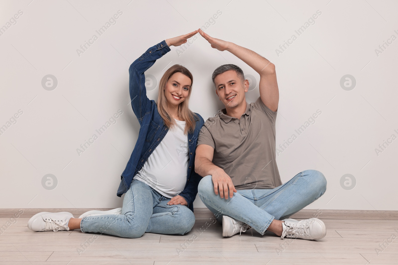 Photo of Young family housing concept. Pregnant woman with her husband forming roof with their hands while sitting on floor indoors