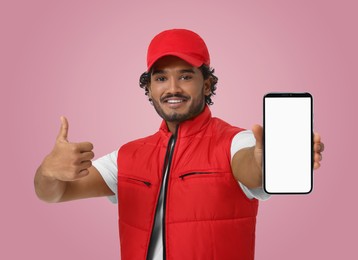 Image of Happy courier holding smartphone with empty screen and showing thumbs up on pink background