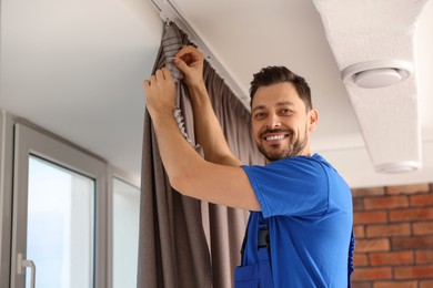 Worker in uniform hanging window curtain indoors