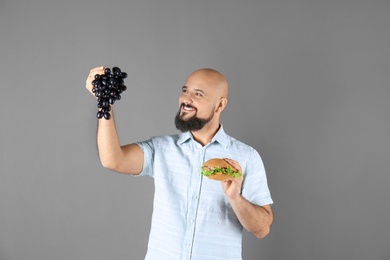Overweight man with hamburger and grapes on gray background