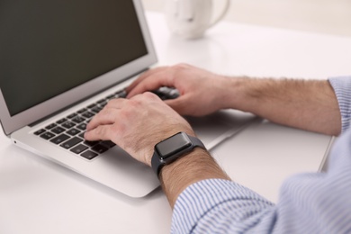 Young man with smart watch working on laptop at table in office, closeup