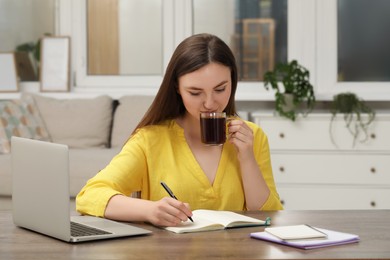 Young woman drinking coffee and writing in notebook at wooden table indoors