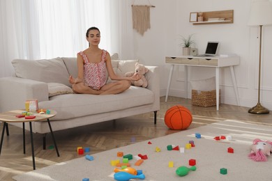 Calm young mother meditating on sofa in messy living room