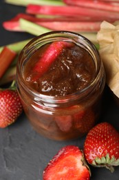 Photo of Jar of tasty rhubarb jam, fresh stems and strawberries on dark textured table, closeup