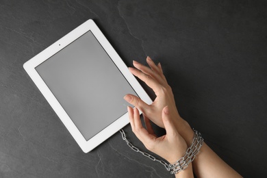 Top view of woman with chained hands and tablet computer on black background, space for text. Loneliness concept