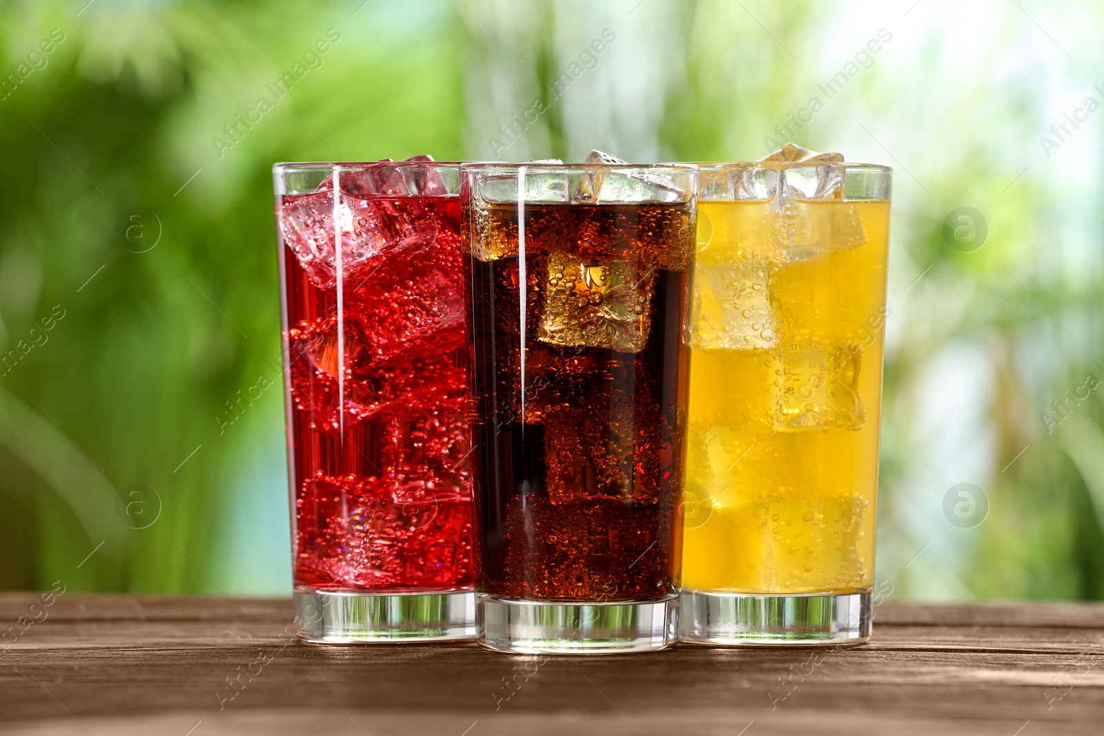 Photo of Glasses of different refreshing soda water with ice cubes on wooden table outdoors