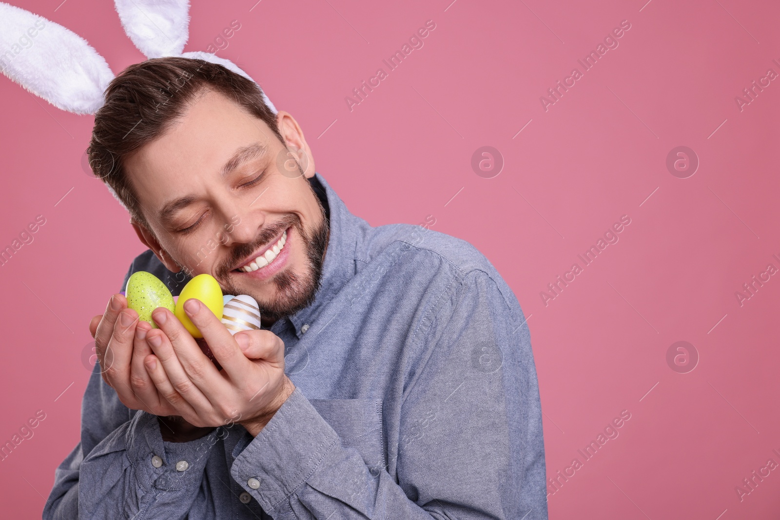 Photo of Happy man in bunny ears headband holding painted Easter eggs on pink background. Space for text