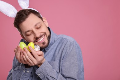 Photo of Happy man in bunny ears headband holding painted Easter eggs on pink background. Space for text