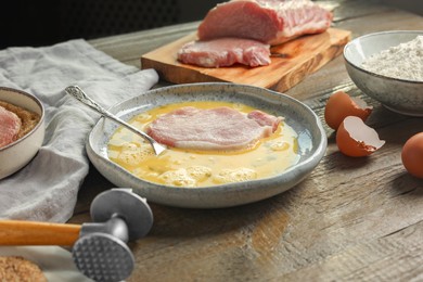 Photo of Cooking schnitzel. Raw pork chop in eggs, meat mallet and ingredients on wooden table